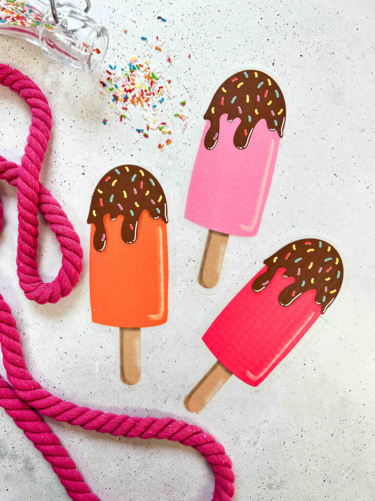 Drei bunte Eis am Stiel Applikationen mit Schokoglasur und Streuseln, arrangiert mit einer pinken Kordel und bunten Zuckerstreuseln auf hellem Grund.
