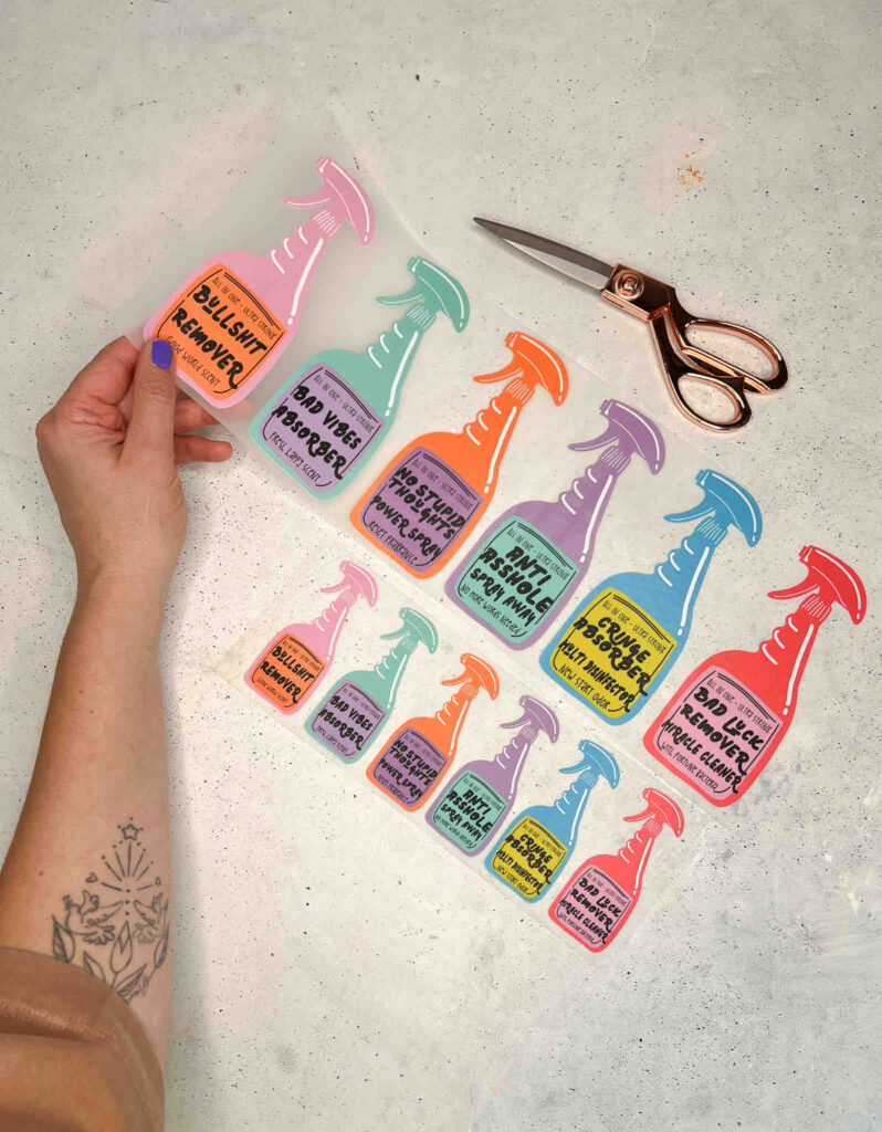 Eine Hand hält einen Bogen mit bunten Sprühflaschen-Aufklebern von Anke, daneben liegen eine Schere und weitere kleine Sticker auf hellem Untergrund.