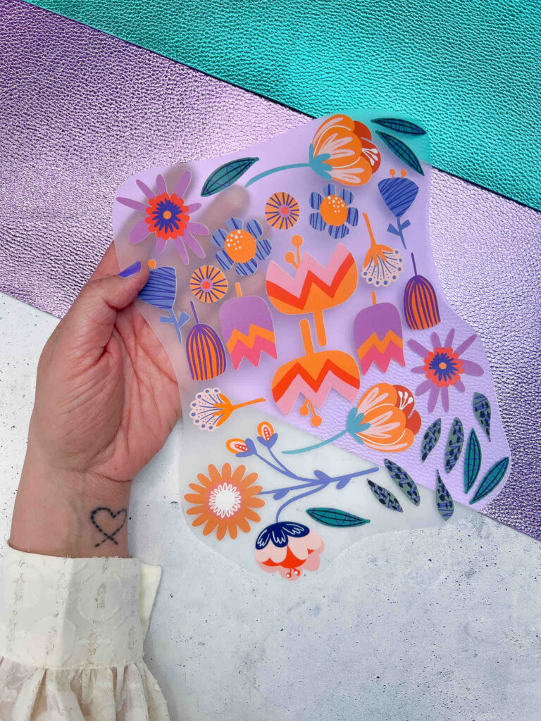 Eine Hand hält eine transparente Folie mit bunten, floralen Illustrationen in Orange, Lila und Blau vor einem glitzernden, violett-türkisen Hintergrund.