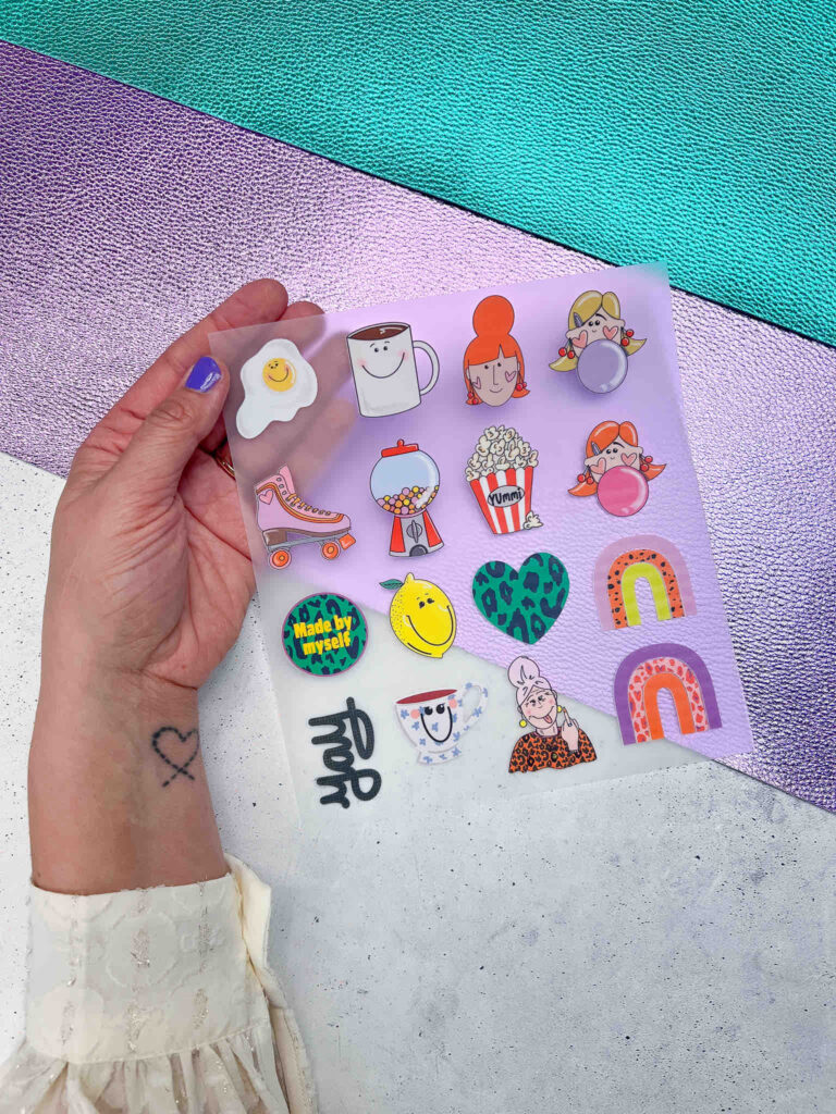 Hand hält das Bügelbild | The Minis #1 mit vielen bunten, verspielten Motiven wie Regenbögen, Herzen, Rollschuhen und Gesichtern auf einer Folie.