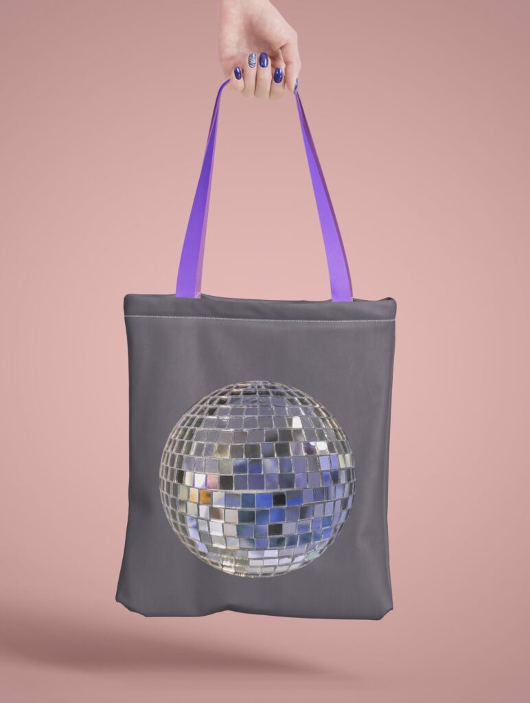 Eine Hand hält eine graue Stofftasche mit violetten Trägern, die mit einer glitzernden Discokugel bedruckt ist, vor einem zartrosa Hintergrund.
