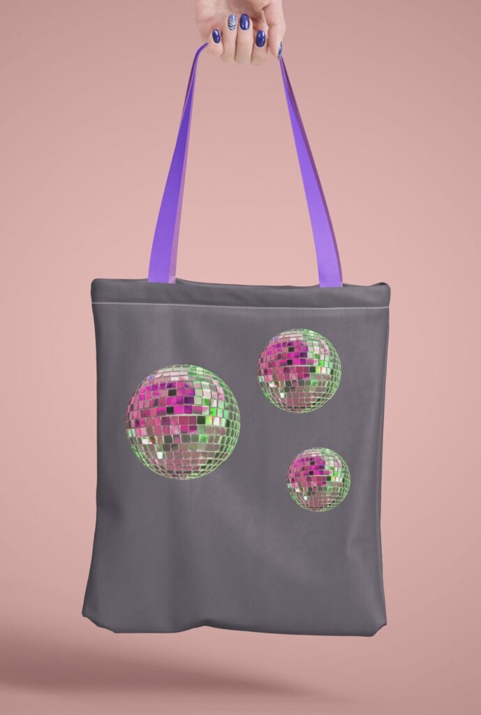 Eine Hand hält eine graue Stofftasche mit violetten Henkeln, die mit drei glitzernden Discokugeln aus dem Bügelbild DISCO Kugel-SET verziert ist.