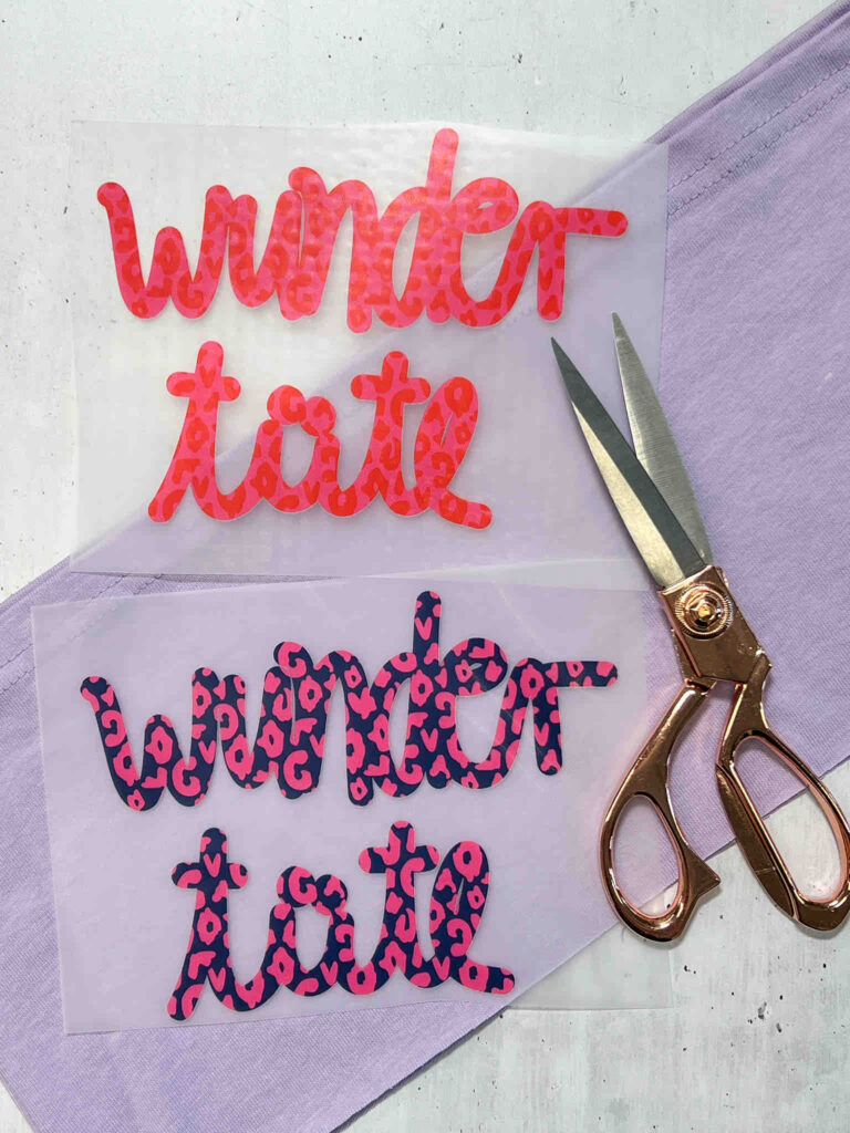 Bügelbilder mit Schriftzug Wundertüte in Leopardenmuster auf Transferfolie neben einer roségoldenen Schere auf einem helllila Stoffuntergrund.