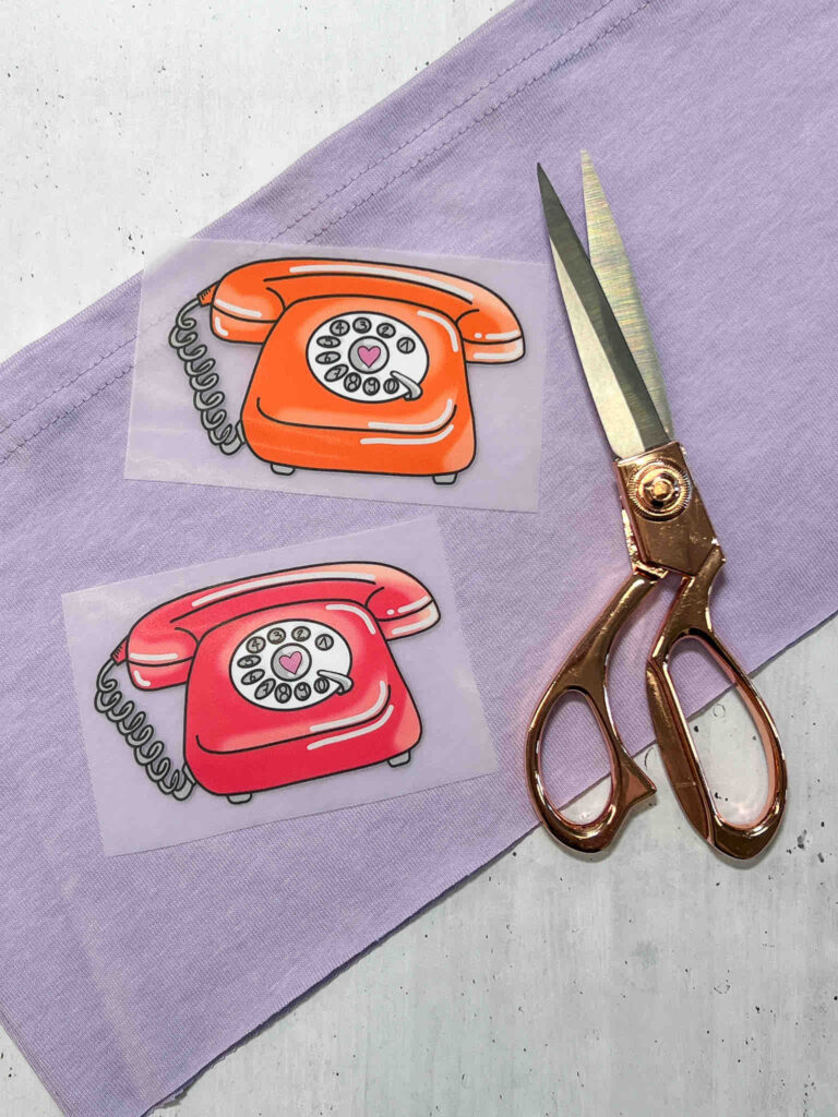 Zwei Aufbügler mit Retro-Telefon-Motiven in Orange und Rot liegen neben einer roségoldenen Stoffschere auf einem helllila Stoffuntergrund.
