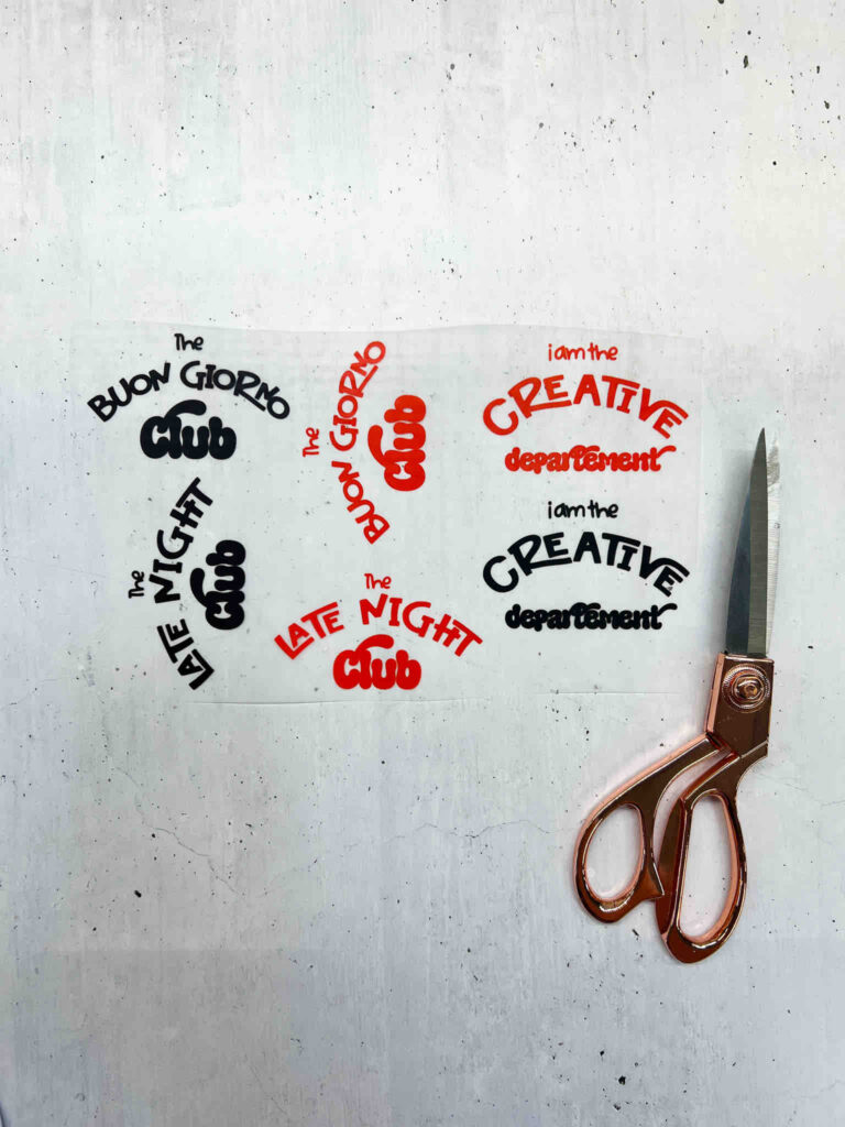 Ein Bogen mit Aufklebern in Rot und Schwarz mit Schriftzügen wie The Creative Departement neben einer roségoldenen Schere auf hellem Untergrund.