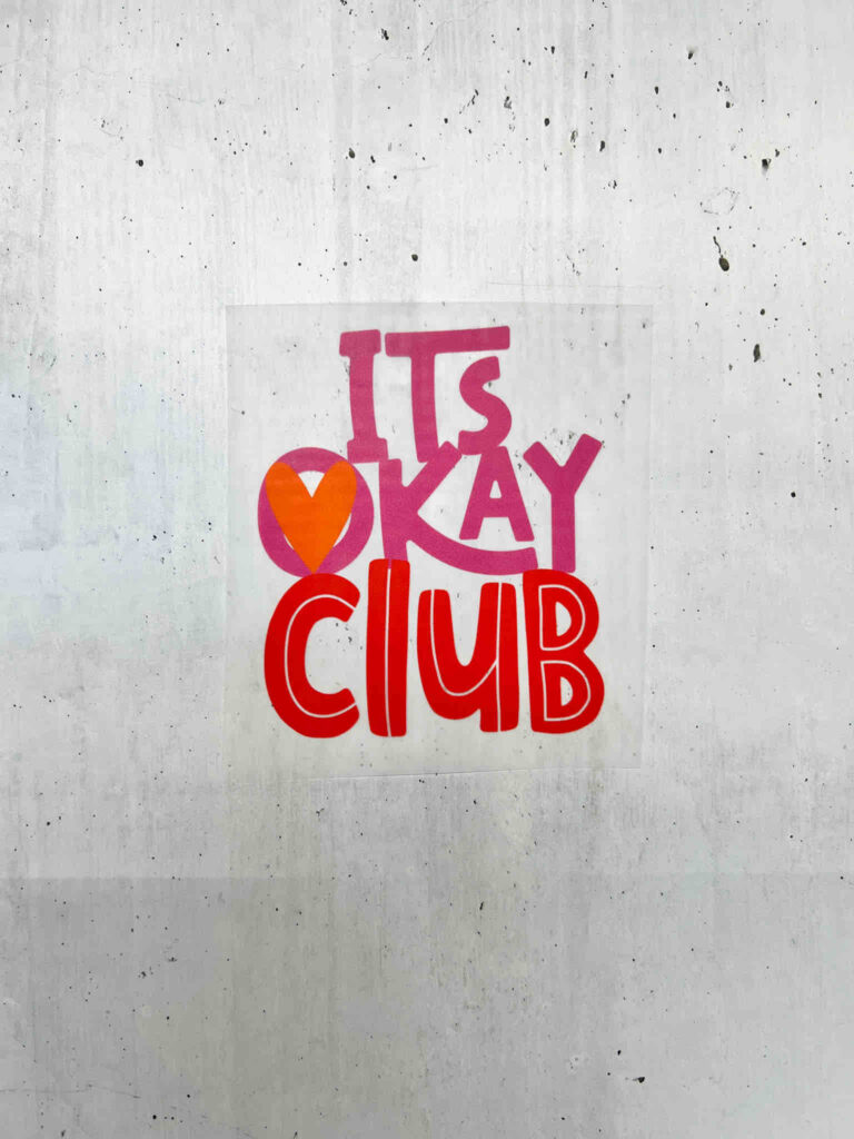 Ein Aufkleber mit der Aufschrift ITS OKAY CLUB in bunten Buchstaben und einem orangefarbenen Herz auf einer hellgrauen, strukturierten Betonwand.
