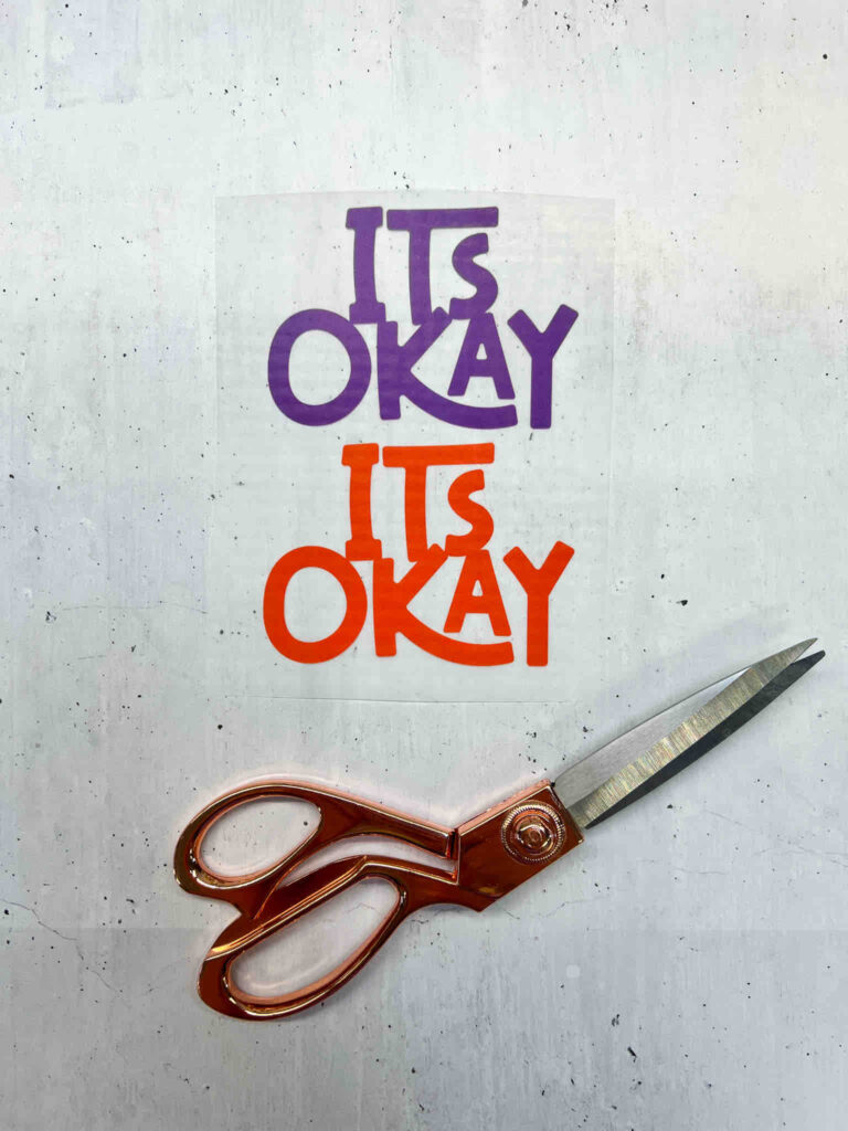 Zwei Aufkleber mit dem Schriftzug „ITS OKAY“ in Lila und Orange auf einer Unterlage, daneben liegt eine hochwertige Schere in Kupferoptik.