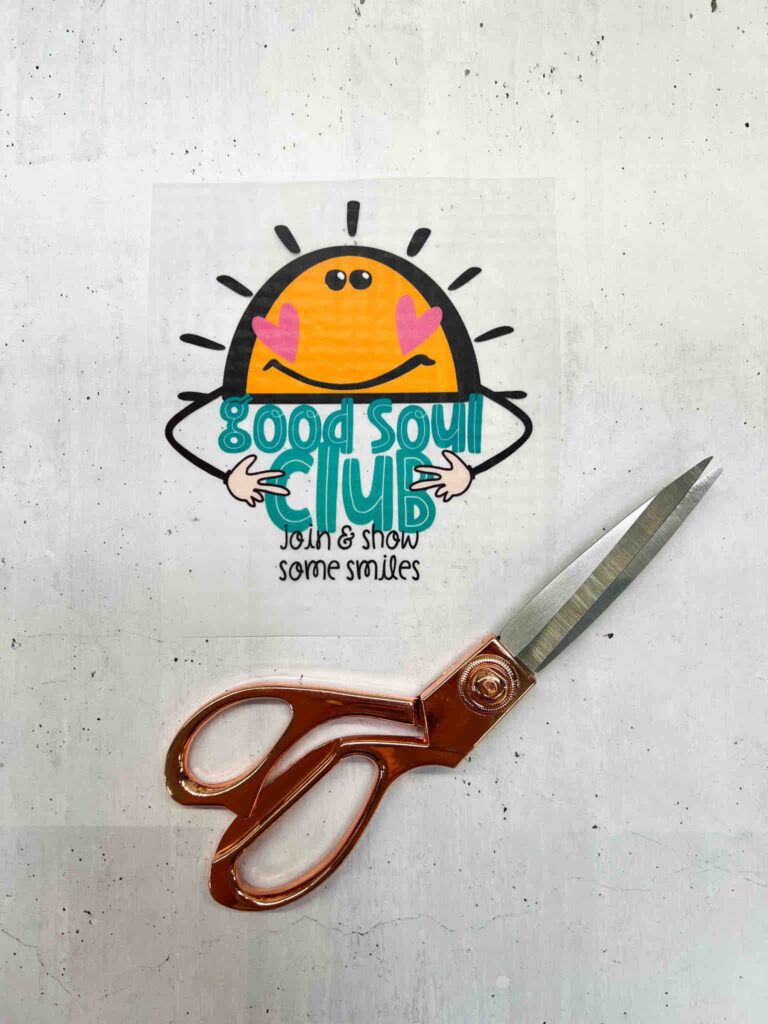 Ein Aufkleber mit einer lächelnden Sonne und dem Schriftzug Good Soul Club neben einer eleganten, kupferfarbenen Schere auf hellem Untergrund.