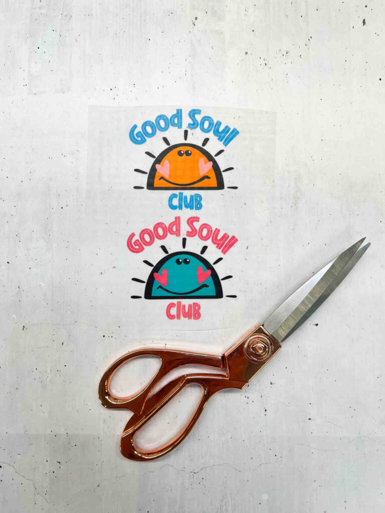 Zwei Good Soul Club Aufkleber mit lachenden Sonnenmotiven in Orange und Türkis neben einer eleganten, kupferfarbenen Schere auf hellem Untergrund.