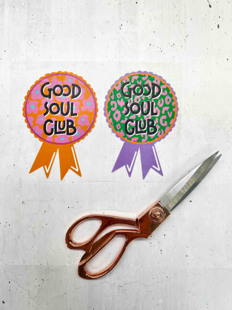 Zwei runde Good Soul Club Aufkleber mit Blumen- und Leopardenmuster neben einer kupferfarbenen Schere auf einem hellen, gesprenkelten Untergrund.