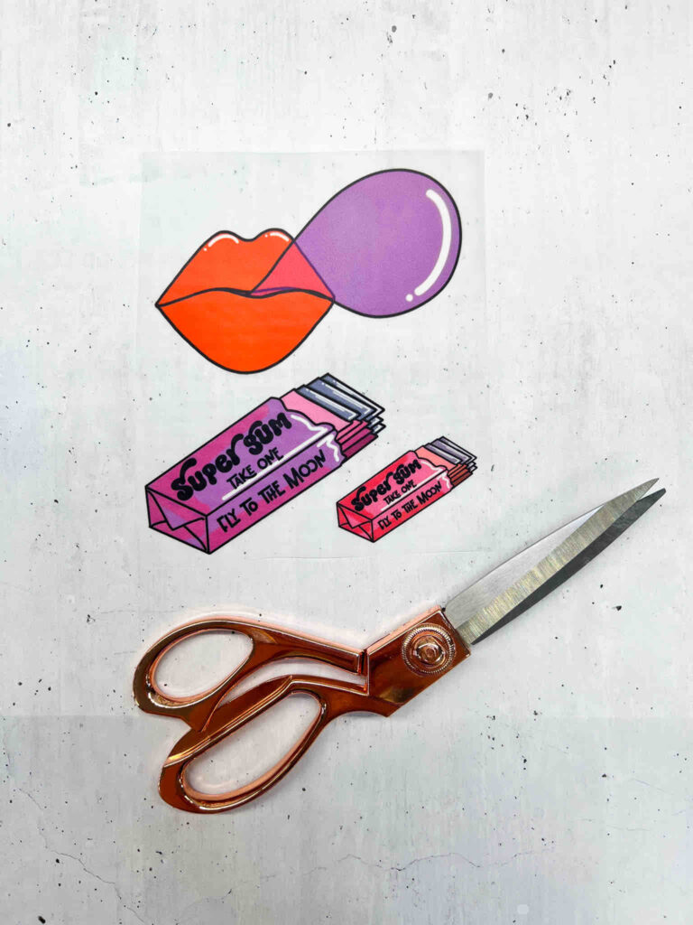 Stickerbogen mit Lippen, Kaugummipackungen und Schriftzug Super Gum neben einer kupferfarbenen Schere auf einem hellen, strukturierten Untergrund.