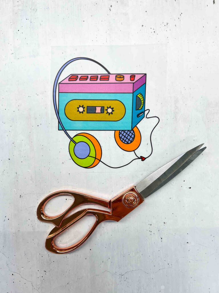 Bügelbild Kassettenspieler mit Kopfhörern im bunten Retro-Design neben einer kupferfarbenen Schere auf einem hellen, gesprenkelten Untergrund.