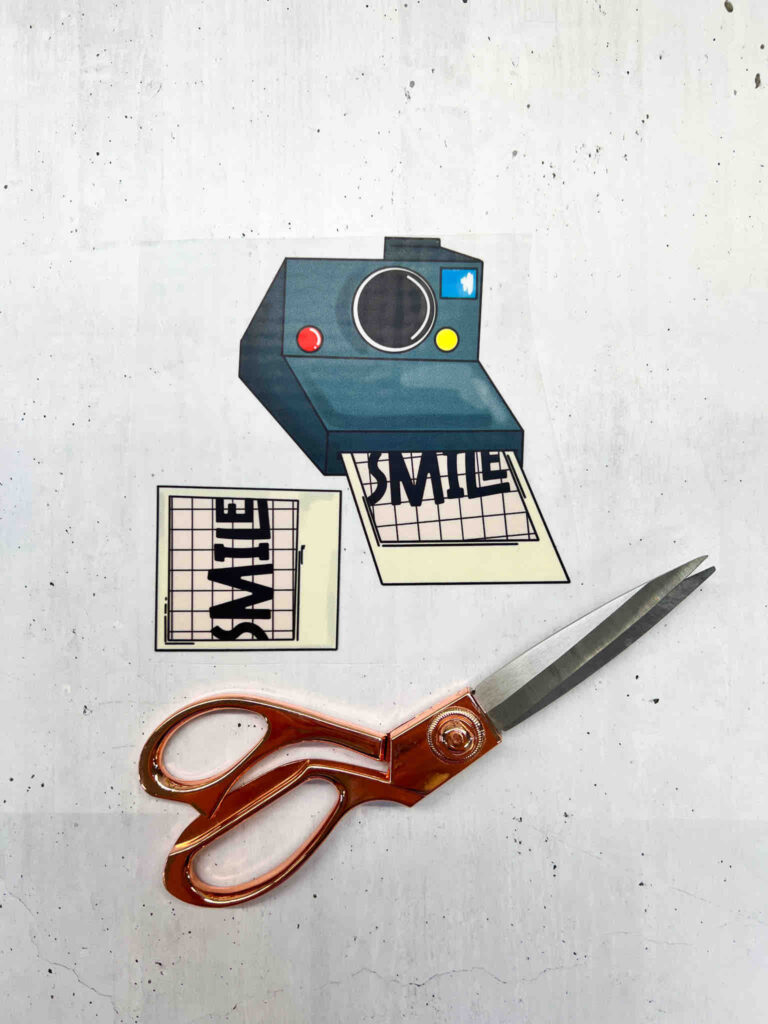 Bügelbild Set mit Sofortbildkamera und Schriftzug SMILE, dazu eine kupferfarbene Schere auf hellem Untergrund für kreative Textilgestaltungen.