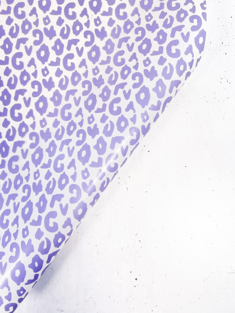 Ein Bogen Geschenkpapier mit einem violetten Leopardenmuster auf weißem Hintergrund, der sich diagonal über einen hellen Untergrund rollt.