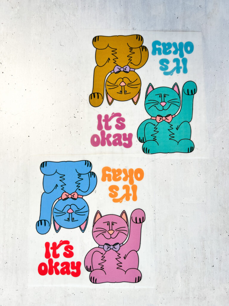 Zwei Bögen mit bunten, illustrierten Katzen-Stickern von Anke, die den Schriftzug „It’s okay“ tragen und in verschiedenen Farben dargestellt sind.