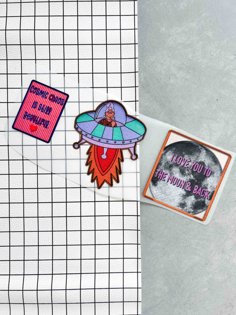 Drei Aufnäher von Anke: ein Schild mit Text, ein UFO mit Figur und ein Mond-Motiv mit Schriftzug, präsentiert auf einem karierten Untergrund.