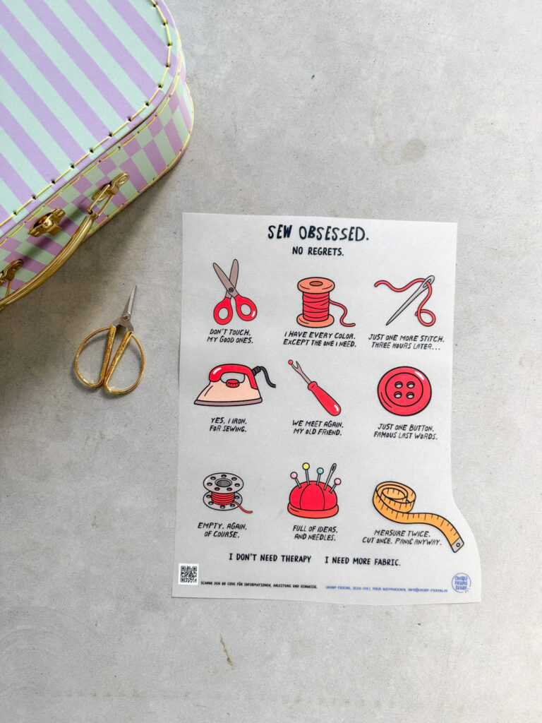 Ein Bügelbild von Cherry Picking mit humorvollen Näh-Illustrationen, daneben eine goldene Schere und ein pastellfarbener Koffer auf grauem Untergrund.