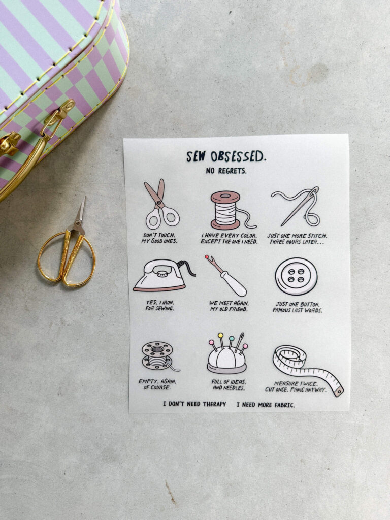 Bügelbild mit Nähutensilien und humorvollen Texten über das Nähen, daneben eine goldene Schere und ein pastellfarbener Koffer auf grauem Untergrund.