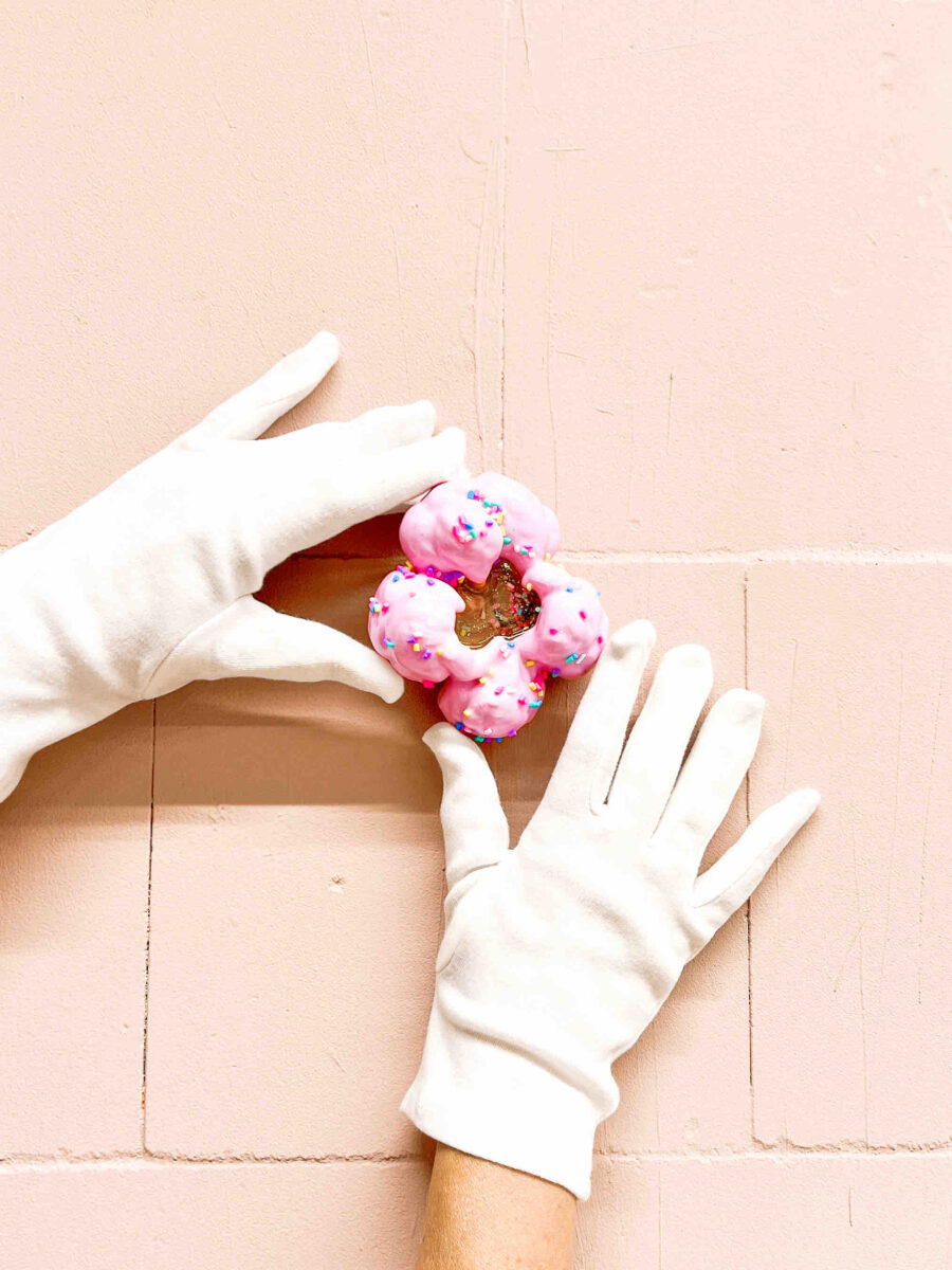 Hände in weißen Handschuhen halten das Pop Art Wandobjekt MELTING POP No. 07 SUGAR PEARLS mit rosa Glasur und bunten Streuseln vor rosafarbener Wand.
