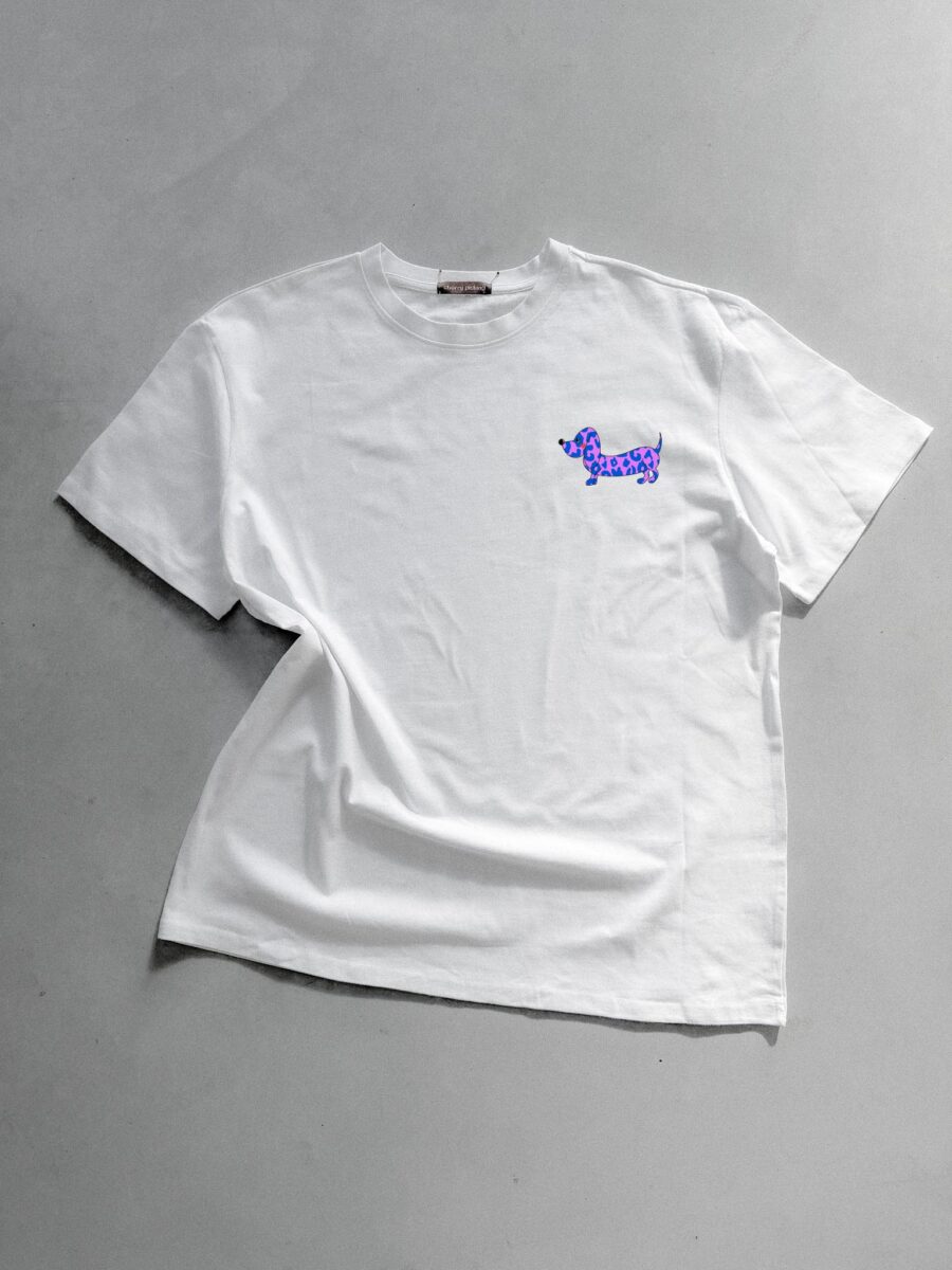 Weißes T-Shirt von Cherry Picking mit einer kleinen, lila-blauen Illustration eines Dackels mit Leopardenmuster auf der Brust vor grauem Hintergrund.