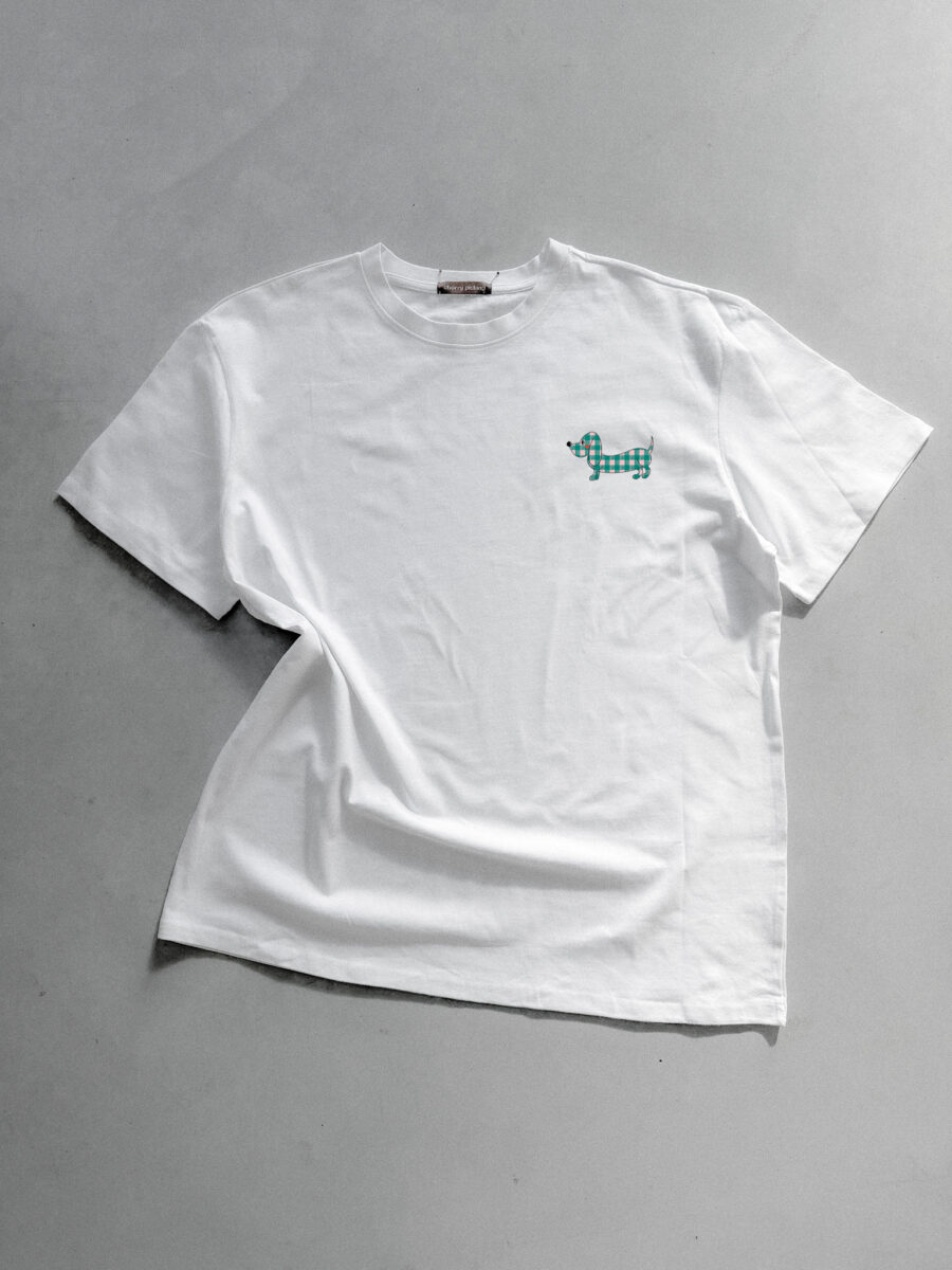 Weißes T-Shirt von Cherry Picking mit einem kleinen, aufgestickten Dackel im grün-weißen Karomuster auf der Brust vor einem hellgrauen Hintergrund.