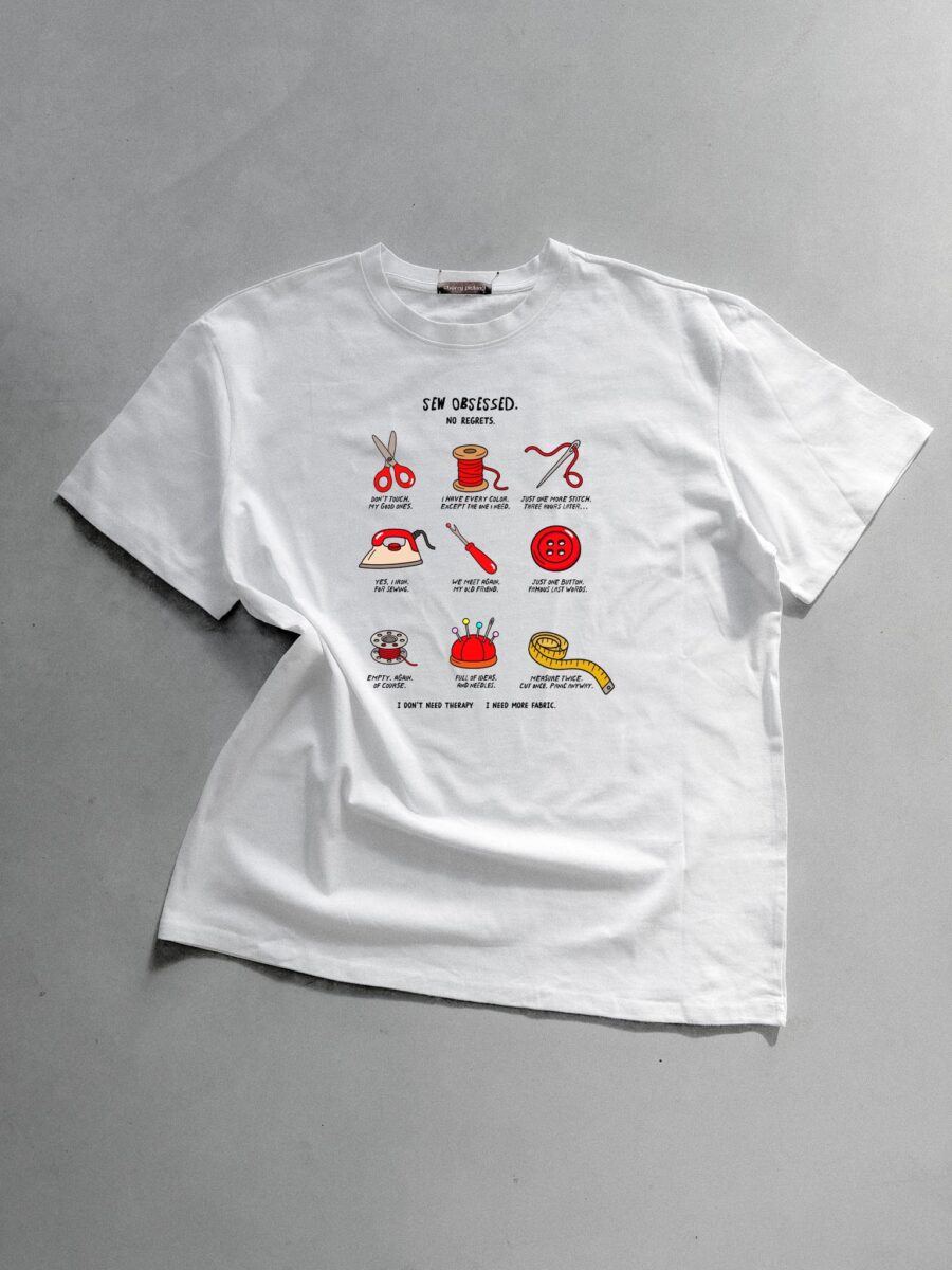 Weißes T-Shirt mit einer Illustration von Nähutensilien und dem Schriftzug Sew Obsessed, präsentiert auf einem grauen Untergrund von Anke.