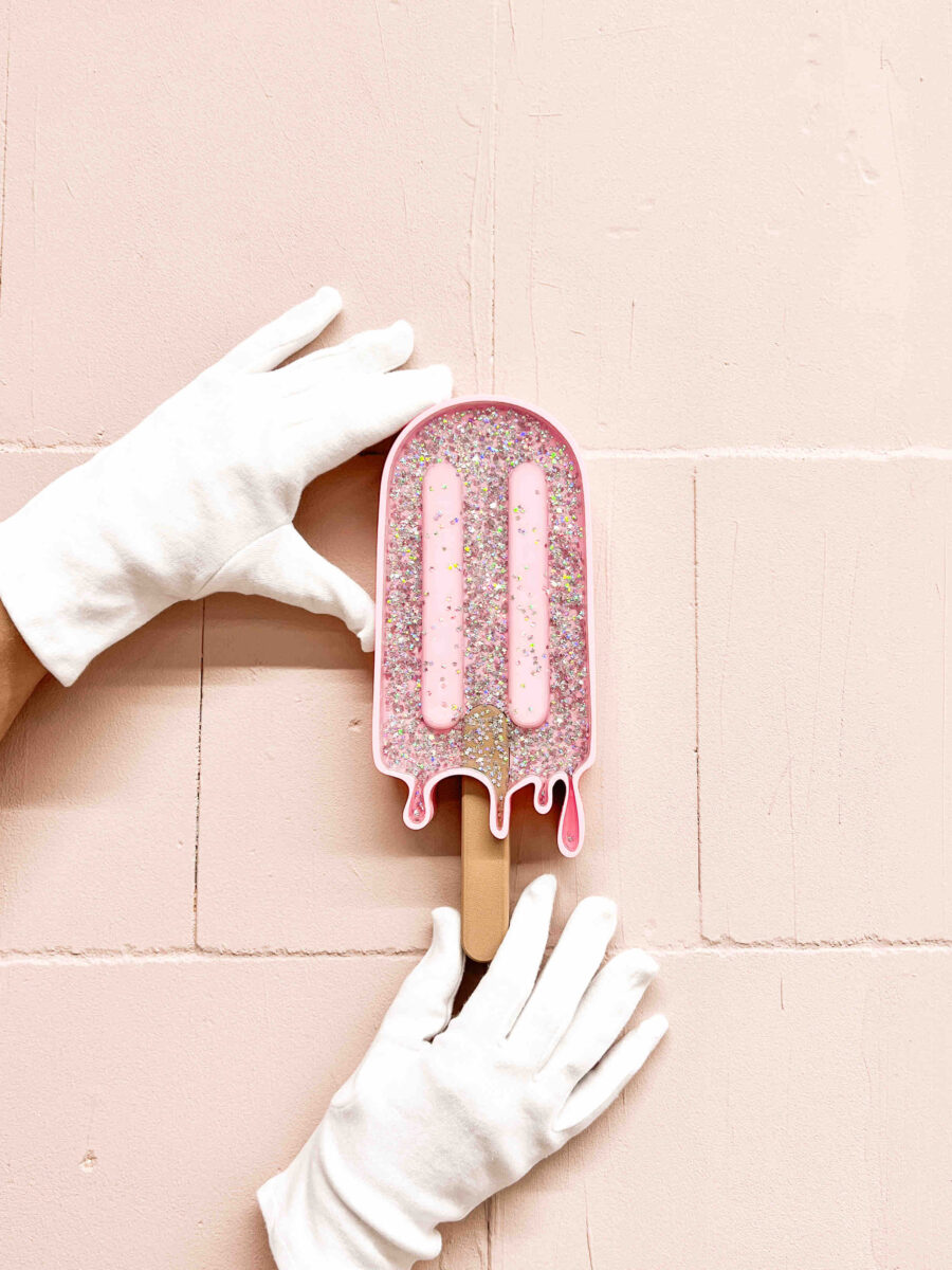 Hände in weißen Handschuhen halten das MELTING POP No. 07 SUGAR PEARLS Wandobjekt, ein glitzerndes, rosafarbenes Eis am Stiel vor einer rosa Wand.