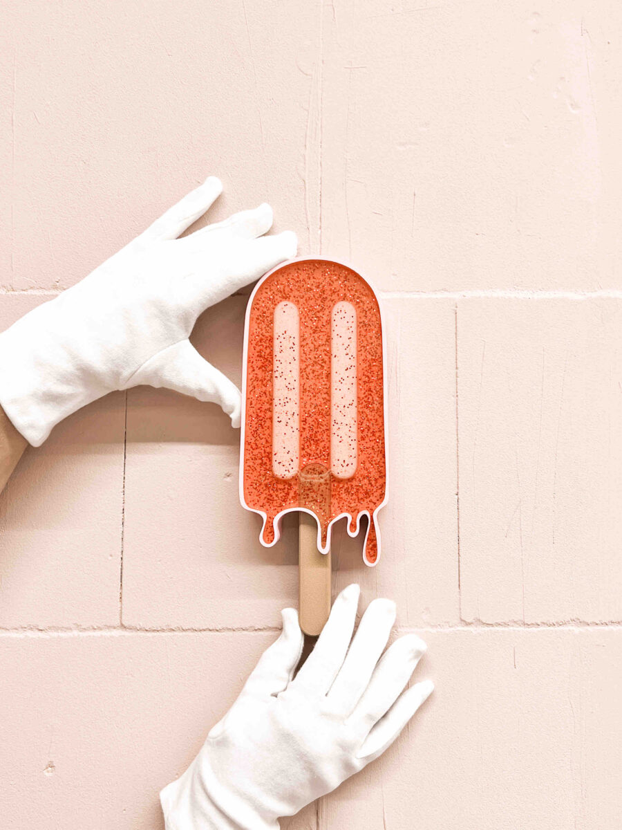 Hände in weißen Handschuhen halten ein glitzerndes, orangefarbenes Eis am Stiel-Objekt von Anke vor einer hellrosa, gekachelten Wand.