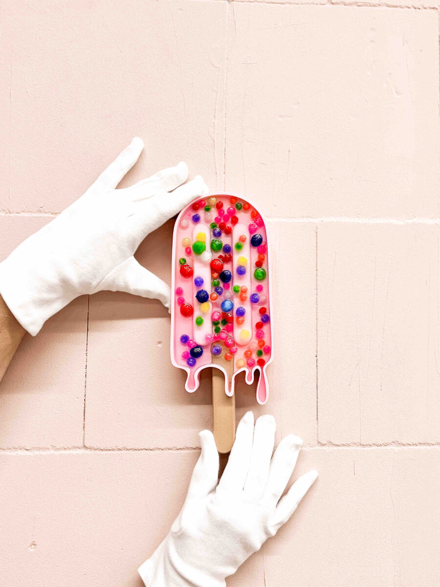 Hände in weißen Handschuhen halten ein rosa Eis am Stiel-Objekt mit bunten, glänzenden Kugeln vor einer hellrosa, gekachelten Wand.
