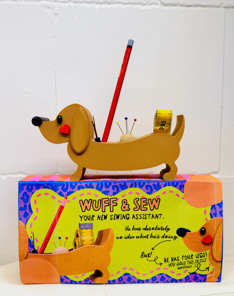 Ein brauner Dackel-Organizer aus 3D Druck als Nähhilfe auf einer bunten Verpackung mit der Aufschrift Wuff & Sew und humorvollem Text über Nähfertigkeiten.