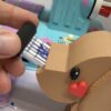 Eine Hand steckt eine Nadelpackung in einen beigen Hundekopf-Halter vor einer hellblauen Regina-Nähmaschine. Ein rotes Herz ziert die Hundeschnauze.