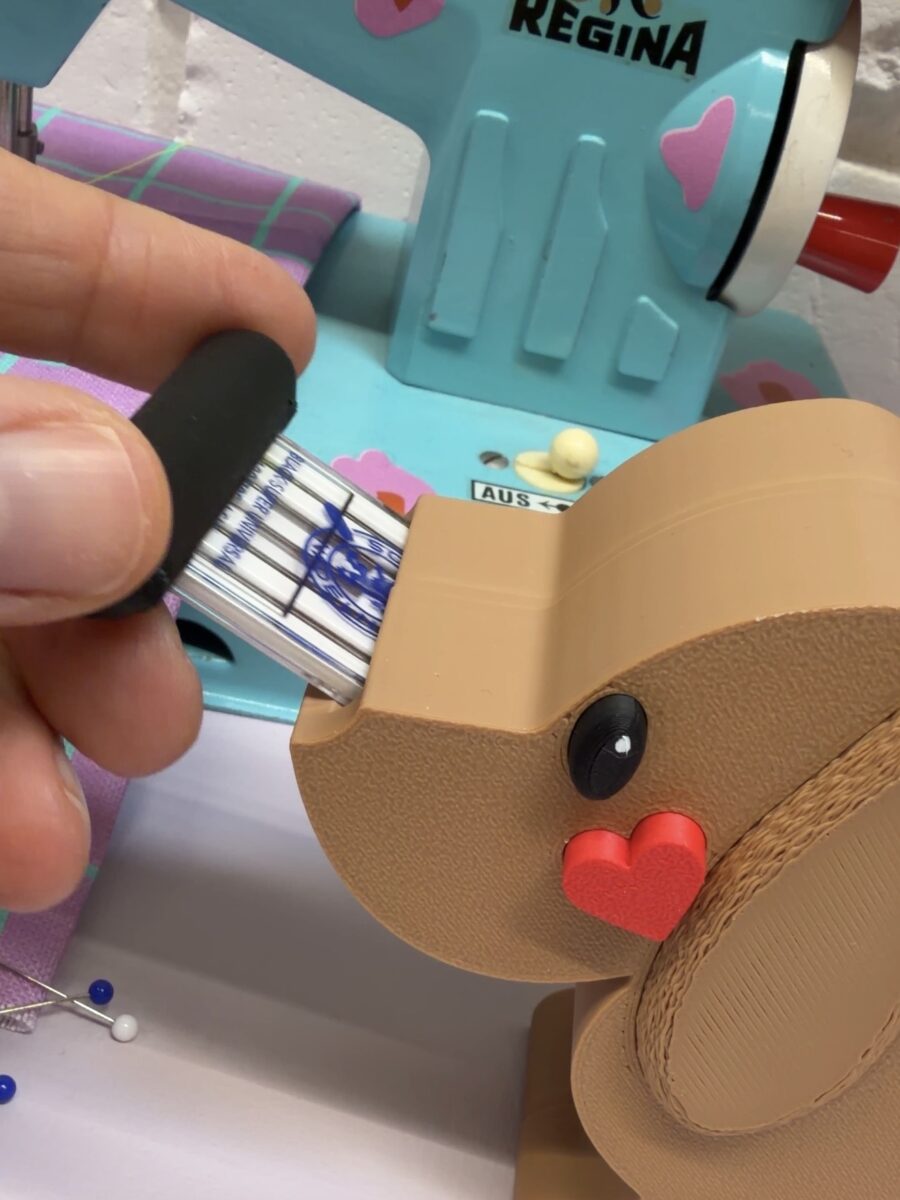 Eine Hand steckt eine Nadelpackung in einen beigen Hundekopf-Halter vor einer hellblauen Regina-Nähmaschine. Ein rotes Herz ziert die Hundeschnauze.