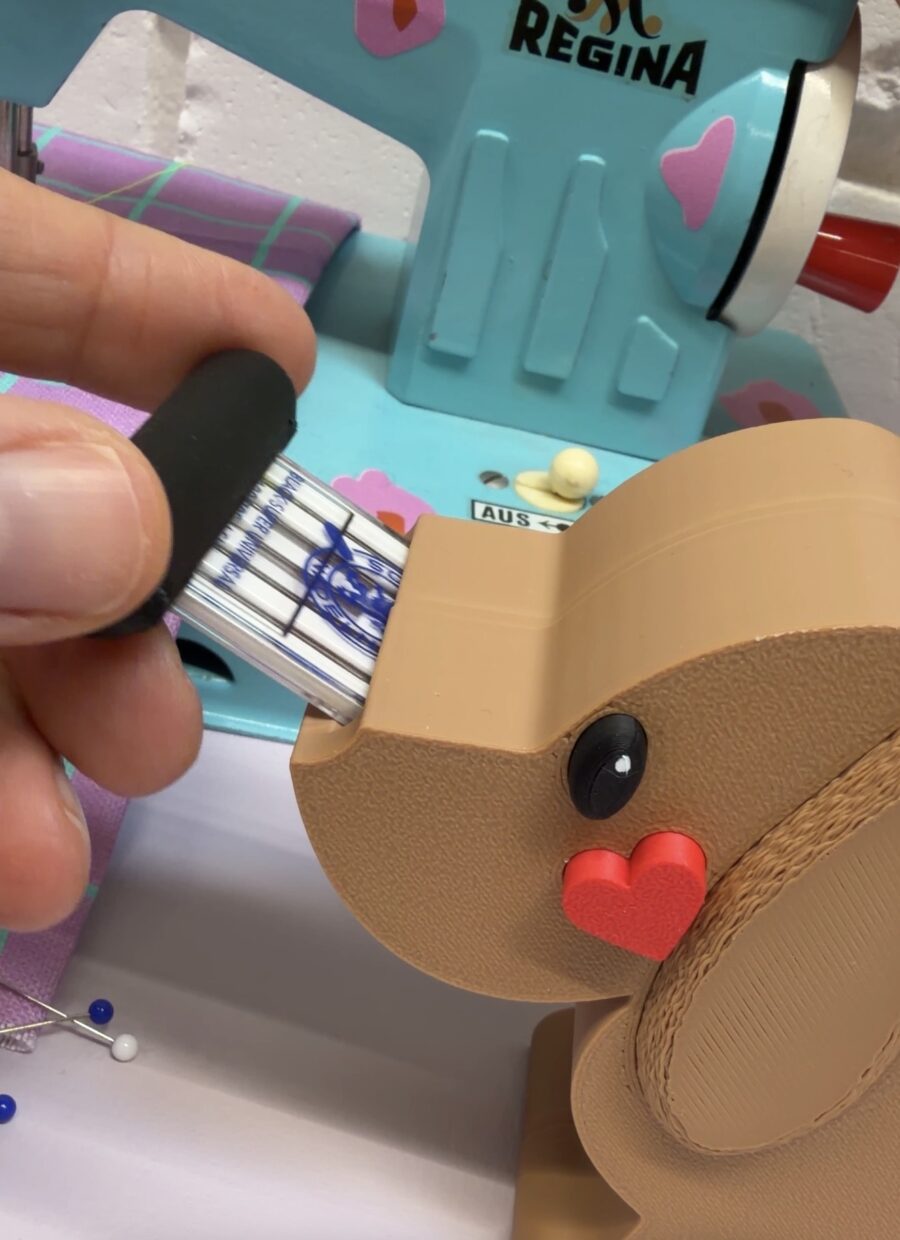 Eine Hand steckt eine Nadelpackung in einen beigen Hundekopf-Halter vor einer hellblauen Regina-Nähmaschine. Ein rotes Herz ziert die Hundeschnauze.